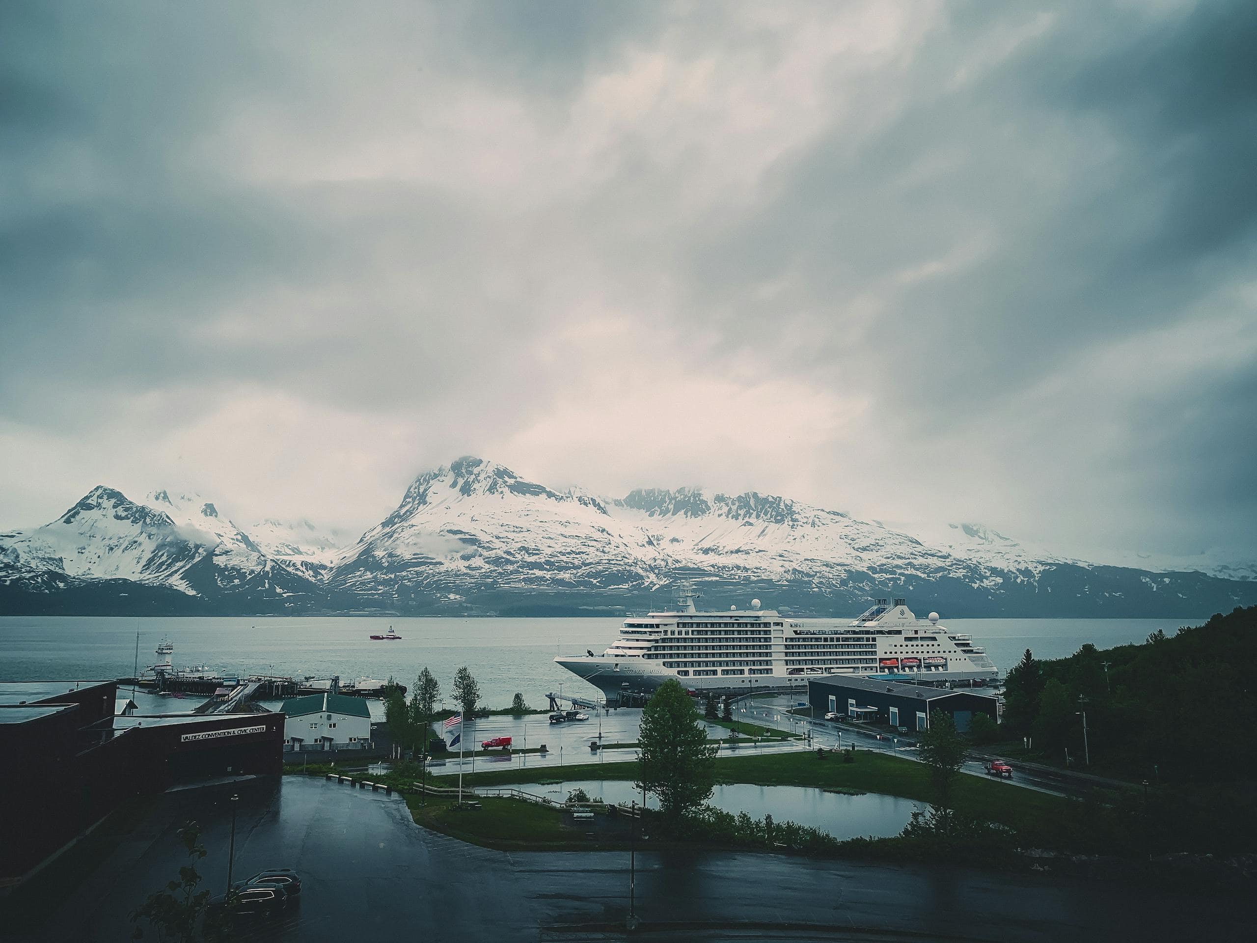 A cruise ship docked with snowy mountains in the background, under a cloudy sky.