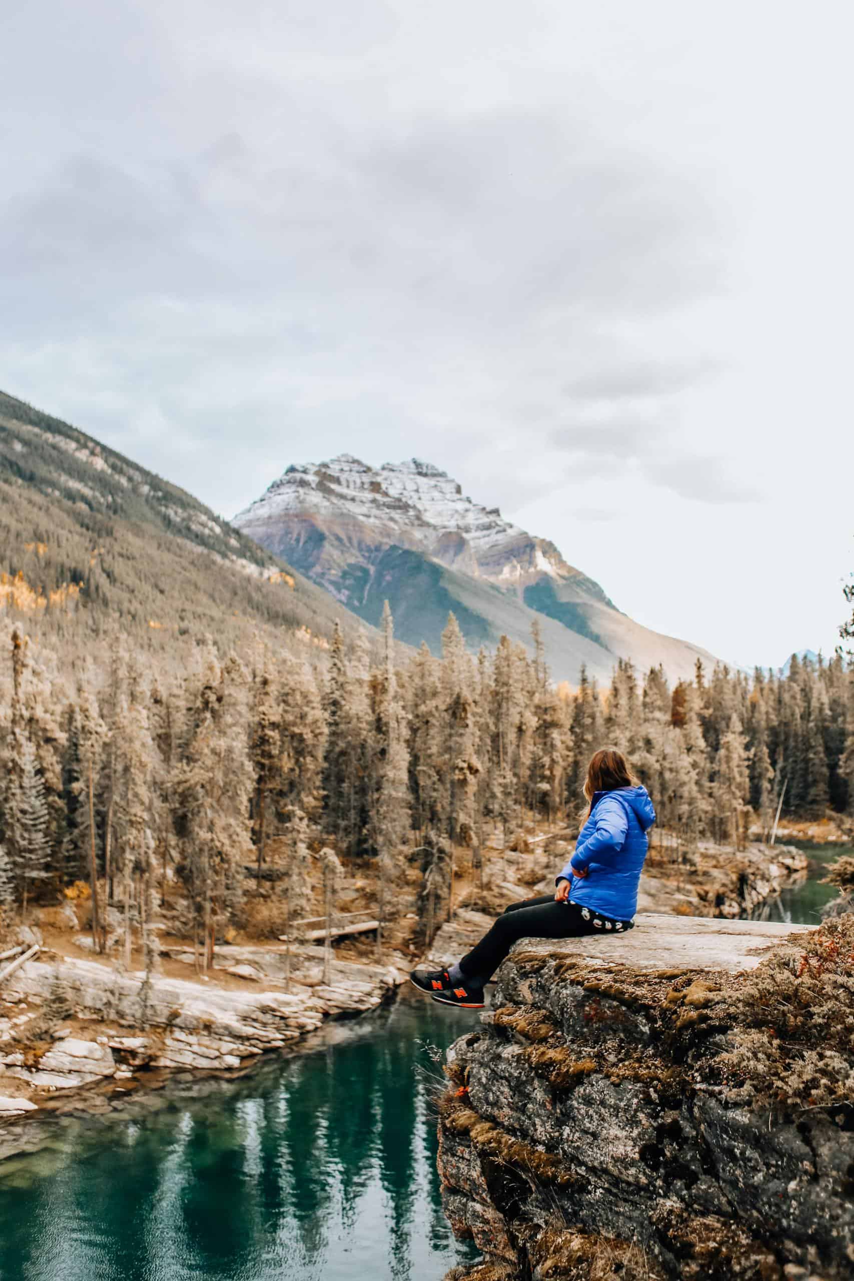 A person in a blue jacket sits on a cliff edge overlooking a river in a mountainous landscape, capturing a sense of adventure.