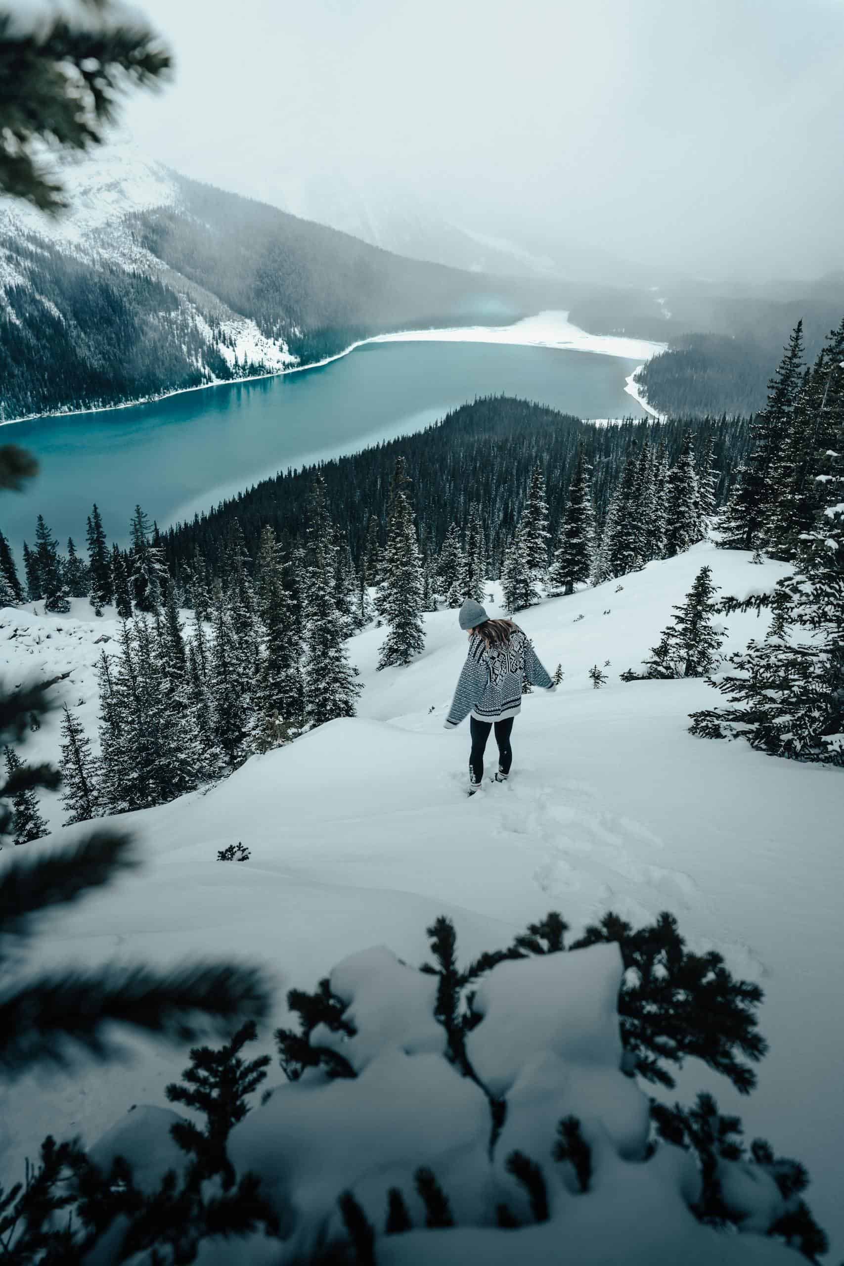 A woman enjoys a snowy hike with a stunning view of a lake and mountain range in Alberta, Canada.