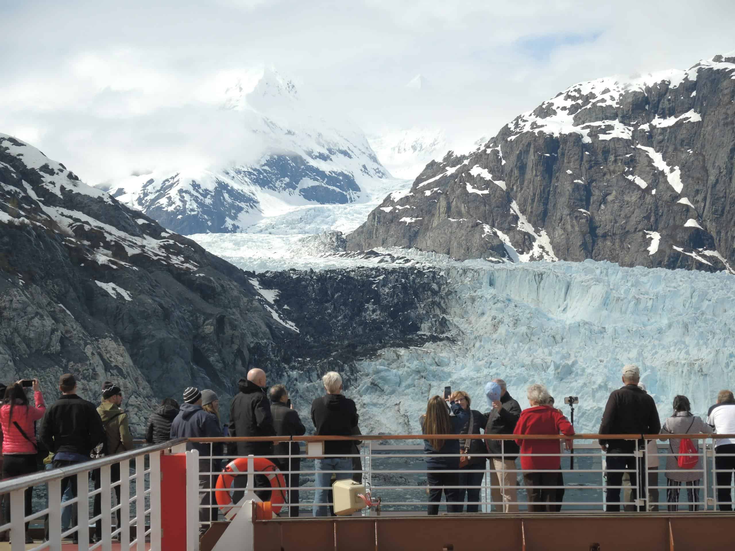 Tourists admire a stunning glacier from a cruise ship deck, surrounded by snowy mountains.